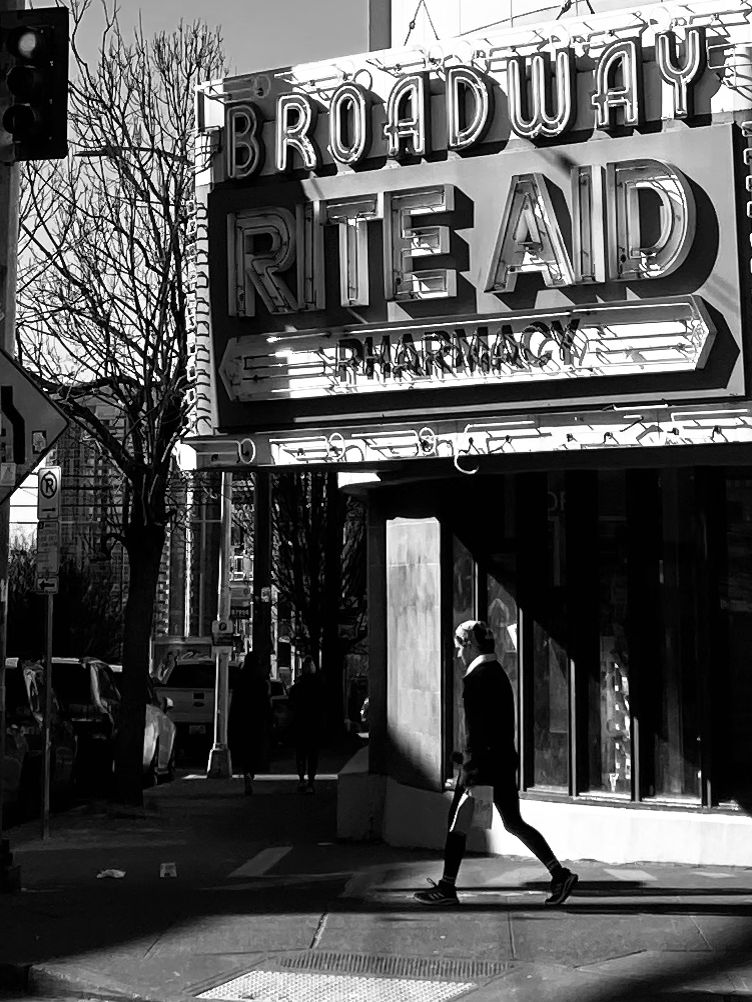 A black and white photo of a person walking in front of the Rite Aid on Broadway, Capitol Hill, Seattle, February 2023.